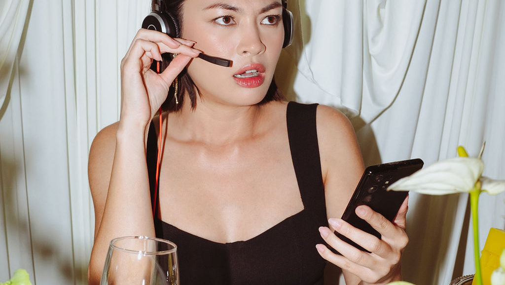 Woman sitting at a table with flowers and food, using a phone and headset.