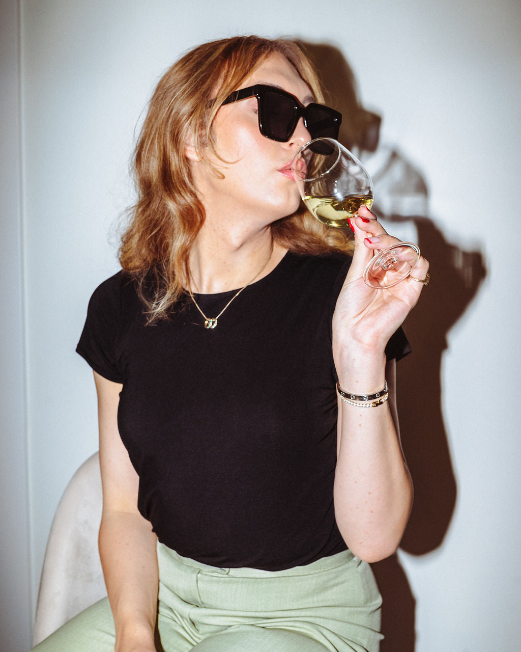 Woman in black top and light green pants drinking from a glass against a white background